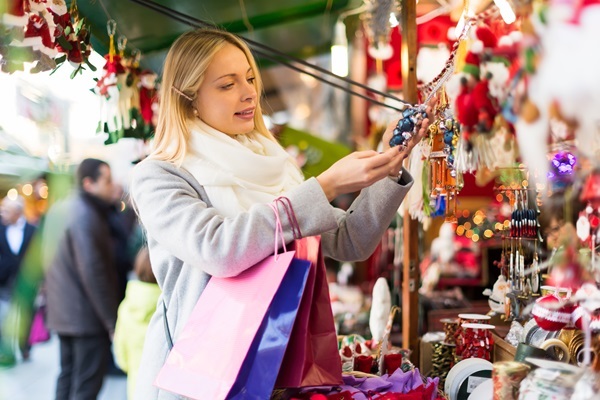 Blonde Frau mit Schal und Einkaufstaschen begutachtet Weihnachtsschmuck an einem Marktstand