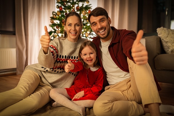 Eltern und kleines Maedchen im roten Kleid sitzen vor Tannenbaum, Eltern zeigen Daumen hoch