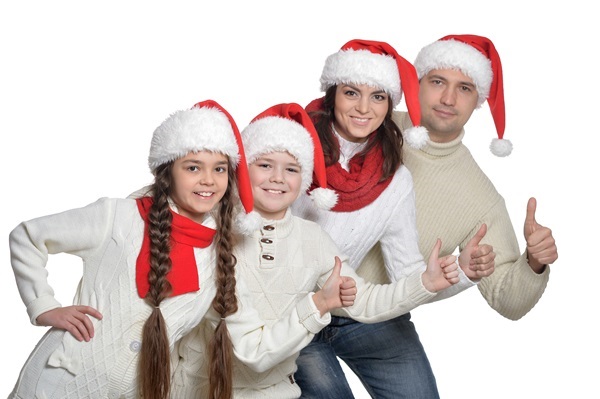 Vierköpfige Familie in weißen Pullovern und roten Weihnachtsmützen steht in Reihe, Vater zeigt Daumen hoch