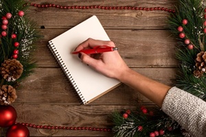 Frauenhand schreibt mit rotem Stift in Notizbuch, umrahmt von Tannenzweigen, Zapfen und Weihnachtskugeln