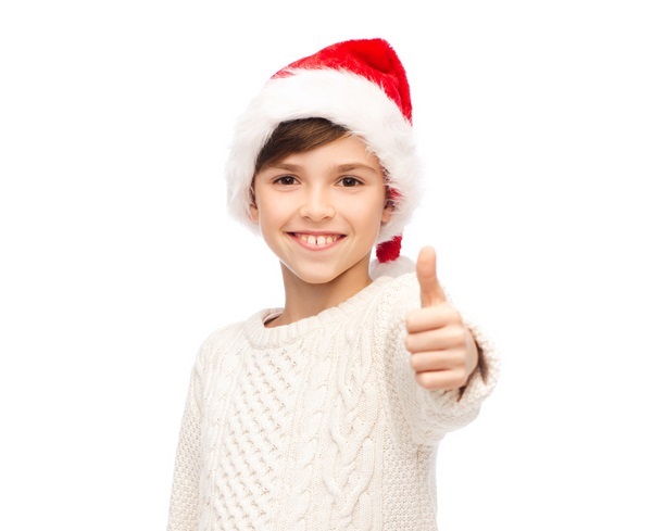 Lachendes Kind mit roter Weihnachtsmütze und weißem Zopfstrickpullover zeigt Daumen hoch
