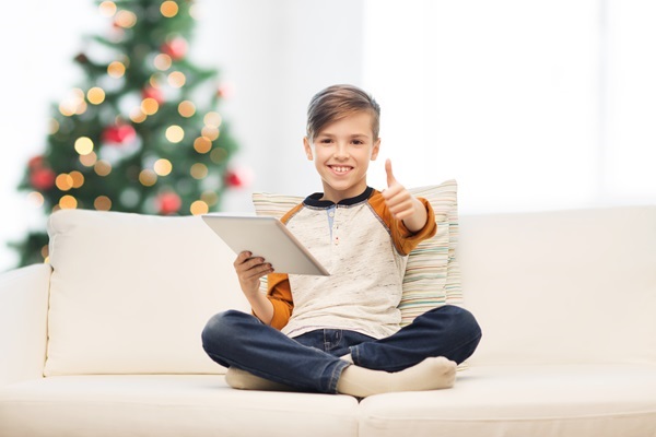 Junge sitzt im Schneidersitz auf Sofa, hält Tablet, zeigt Daumen hoch, Weihnachtsbaum dahinter