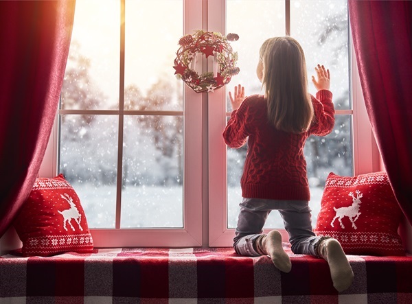Mädchen im roten Pullover kniet auf Fensterbank und schaut nach dem Rentierschlitten, Weihnachtskranz am Fenster