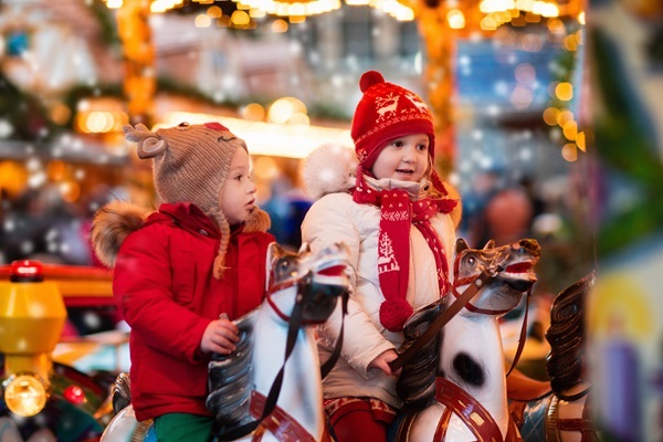 Zwei Kinder in roten Winterjacken auf Karussellpferden auf einem Weihnachtsmarkt mit Lichterketten