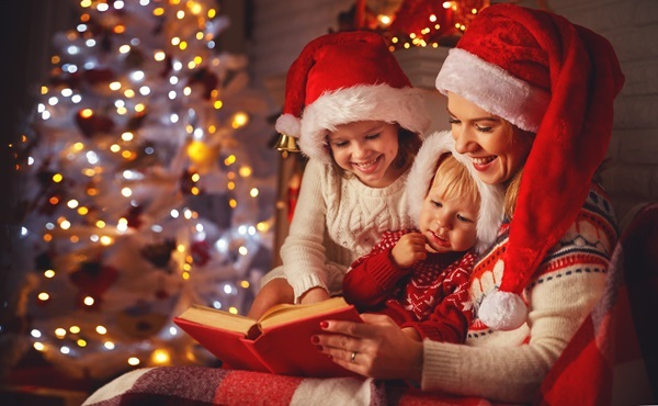 Mutter und Kinder mit Weihnachtsmützen liest zwei Kindern aus rotem Buch vor, Weihnachtsbaum und Kamin dahinter