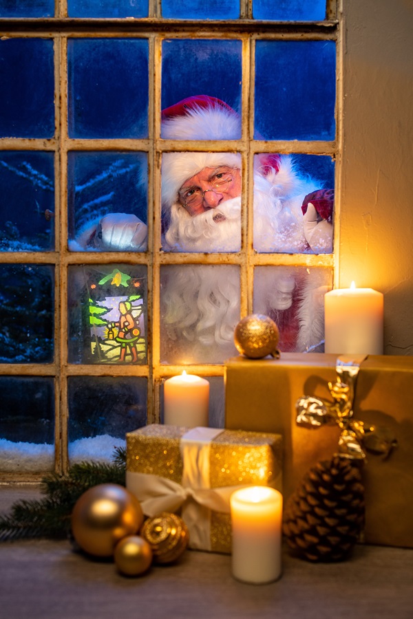 Weihnachtsmann Claudius schaut durch altes Sprossenfenster herein, Kerzen, Teddybär und Geschenke davor, Tannen im Schnee dahinter