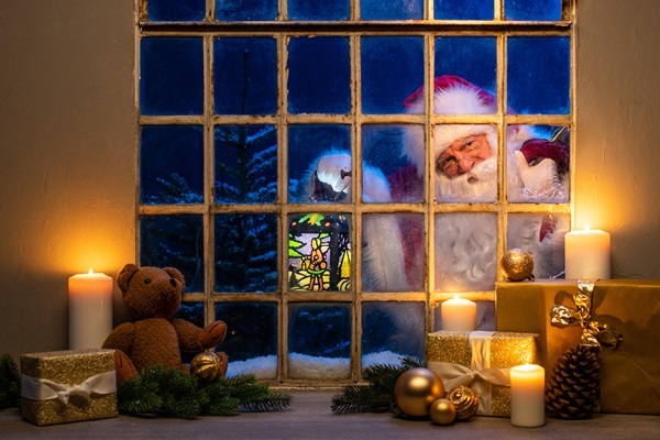 Weihnachtsmann Claudius schaut durch altes Sprossenfenster herein, davor Kerzen, Teddybär und Geschenke, Tannen im Schnee dahinter