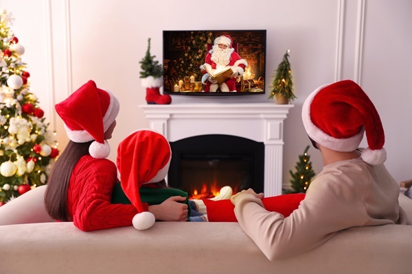 Familie mit Weihnachtsmützen schaut auf einem Fernseher über dem Kamin eine Videobotschaft von Weihnachtsmann Claudius