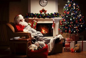 Ein Weihnachtsmann im Ohrensessel vor dem weißen Kamin, Decke über den Füßen, daneben ein Weihnachtsbaum, hübsch dekoriert