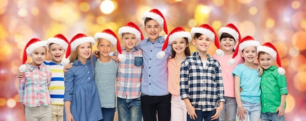 Zehn Kinder mit roten Weihnachtsmützen stehen lachend und sich umarmend vor orangefarbenem Hintergrund