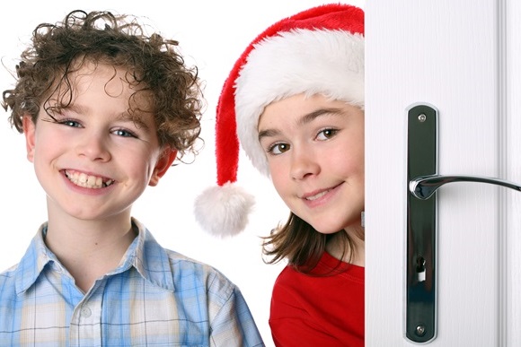 Lachender Junge im Karohemd sowie Mädchen mit roter Weihnachtsmütze lugen neugierig hinter weißer Tür hervor